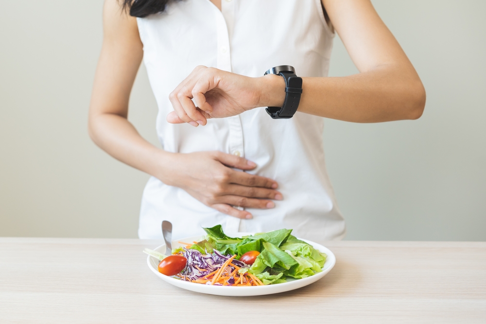 Intermittent fasting, health care asian young woman dietary, having stomach ache, temptation hungry of breakfast food in morning on table at home, looking at watch on her wrist but not yet time to eat