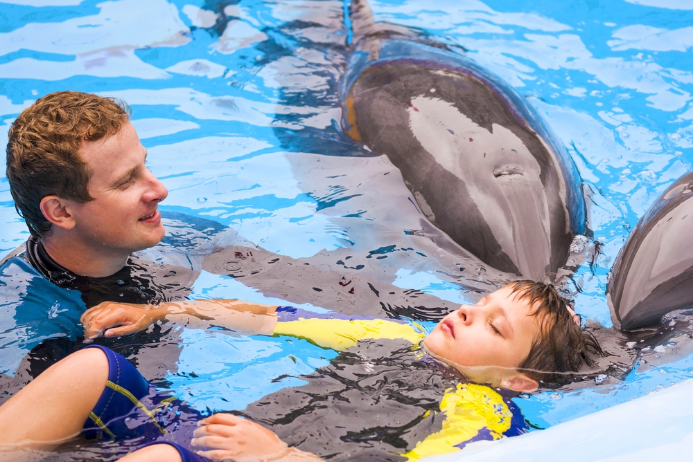 Trainer with boy and dolphin. Treatment of children by means of dolphins. Children and trainer in water petting dolphin. People and dolphins. Concept Dolphin therapy. Kharkiv. Ukraine. 7.15.2018