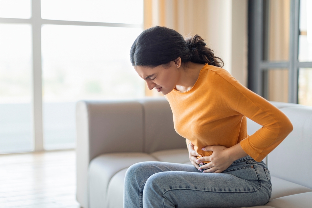 Stressed young indian woman touching her stomach, eastern female having abdominal pain or cramps, sitting on couch with pained expression on her face, frowning while feeling unwell, copy space