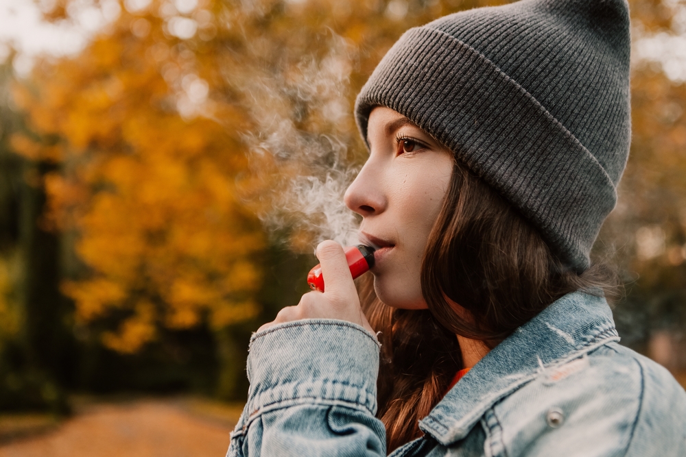 Young woman outside autumn park fall and smoking tobacco device electronic cigarette heater. Smoke and steam system with sticks inside, image with copy space. Harmful habit harm to health lungs