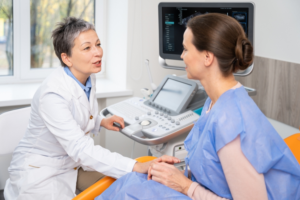 A doctor and patient converse during an ultrasound appointment, with the doctor operating the machine and the patient in a blue gown, seated comfortably.
