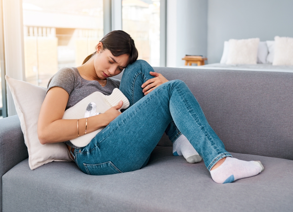 Stomach pain, stress or girl on sofa with hot water bottle for pms gas, constipation or period crisis at home. Tummy, digestion or anxiety in living room with heating pad for endometriosis or pcos