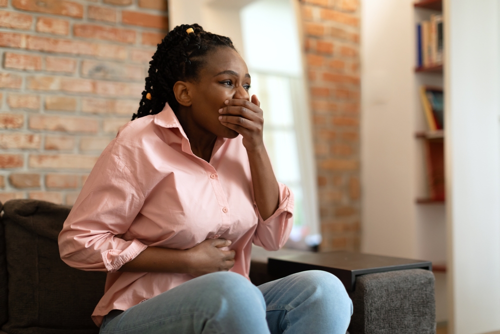 A young black woman sits on a gray couch in a living room with exposed brick, her hand over her mouth as if she is suppressing nausea. Her other hand rests on her stomach, copy space