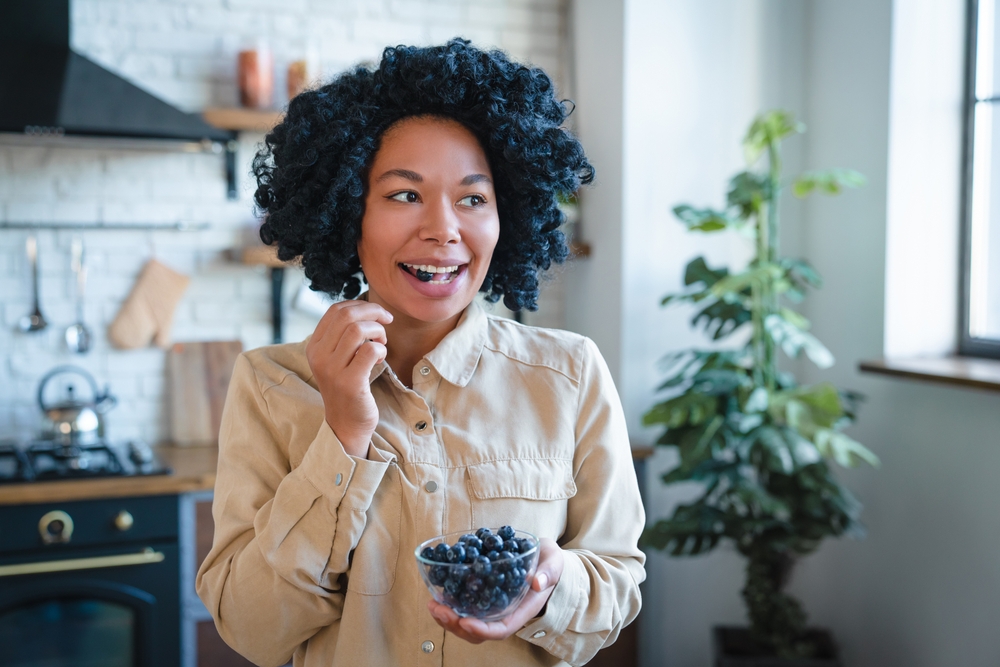 Photo of young smiling healthy laughing african woman in casual eating breakfast and fruits berries blueberries in modern light kitchen in the morning