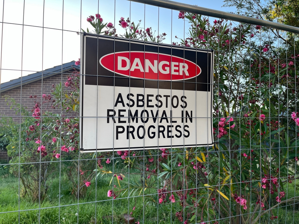 Melbourne, Victoria, Australia - 6 January 2025: A fenced-off area with a prominent "Danger: Asbestos Removal in Progress" sign. The scene includes blooming pink flowers and a residential building.