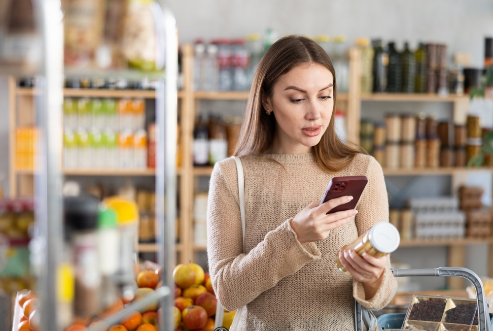 Interested young woman making purchases in local grocery store, using convenient mobile app to pay online for pickled asparagus by scanning barcode on jar