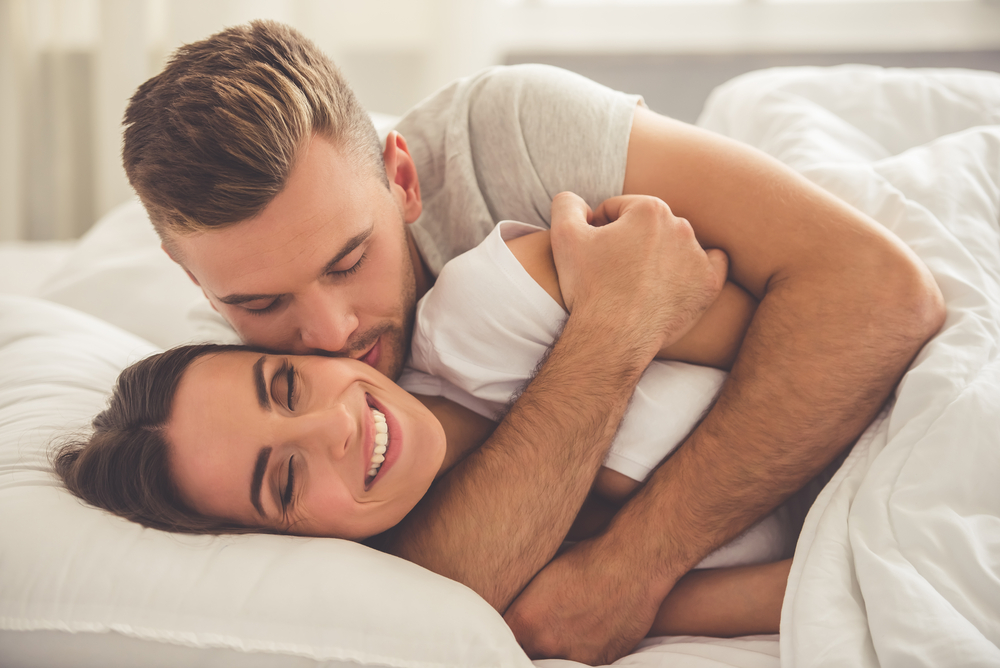Handsome young man is hugging and kissing his beautiful smiling wife in cheek while they both are lying in bed in the morning