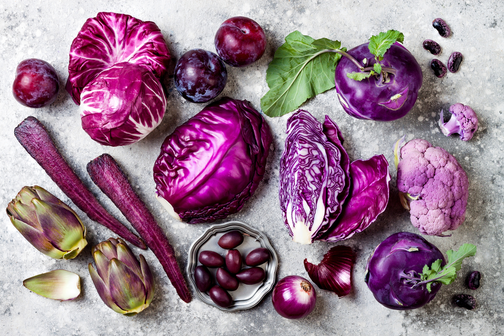 Raw purple vegetables over gray concrete background. Cabbage, radicchio salad, olives, kohlrabi, carrot, cauliflower, onions, artichoke, beans, potato, plums. Top view, flat lay.