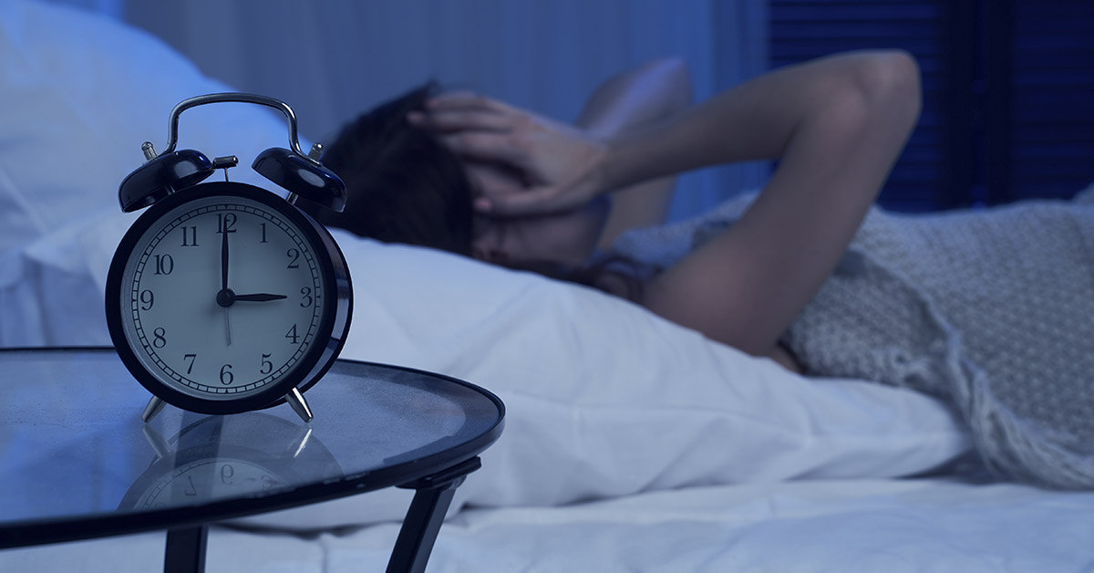 person sleeping in bed, with an alarm clock on the nightstand beside them. The clock is a traditional analog style, and the room appears to be dimly lit, suggesting it is nighttime