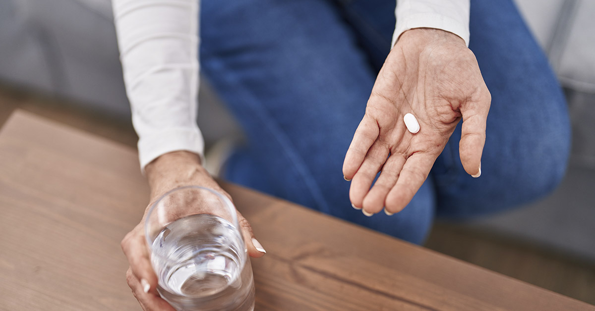 woman holding glass of water in one hand and a white pill in the other