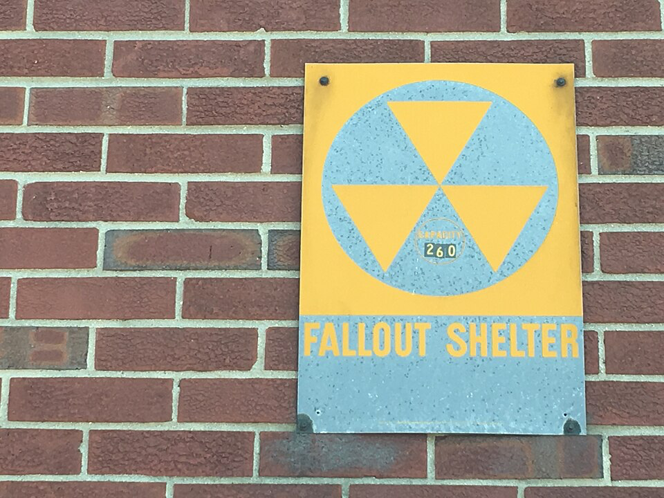 Yellow and blue fallout shelter sign with radiation symbol mounted on brick wall.