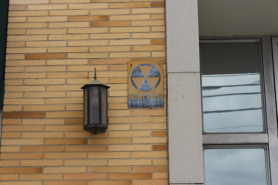 Weathered fallout shelter sign with radiation symbol on exterior brick building wall.