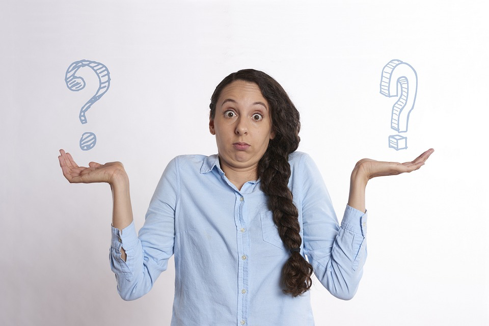 Woman in blue shirt with confused expression holding up both hands beneath question marks, illustrating the paralyzing indecision that prevents people from discarding items.