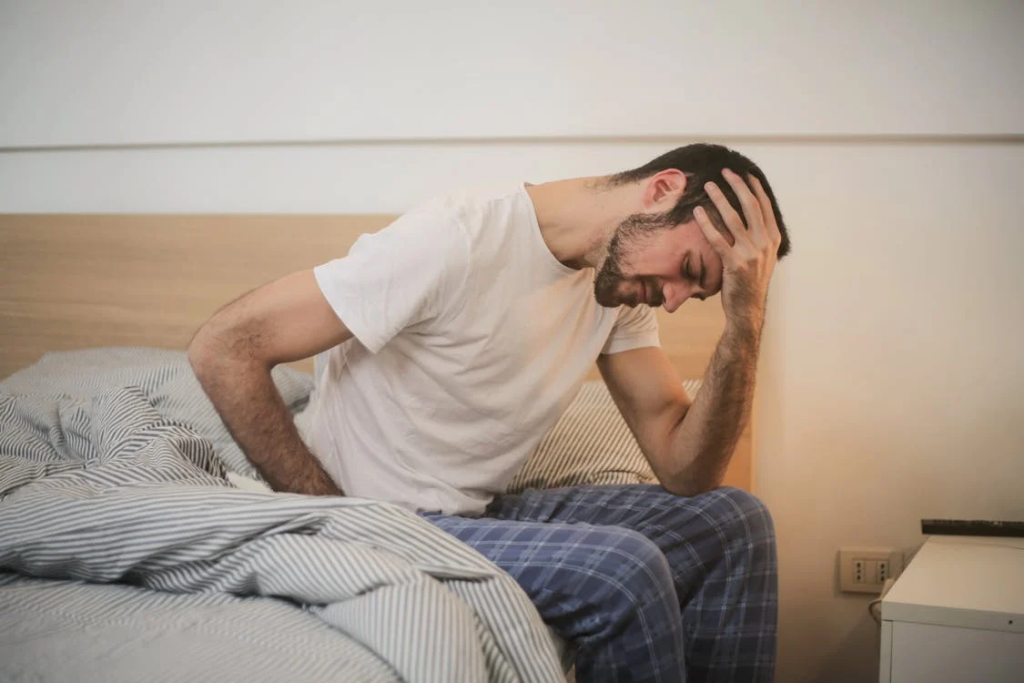 Man sitting on bed edge holding his head in his hands, showing cancer symptoms at night including exhaustion and distress.