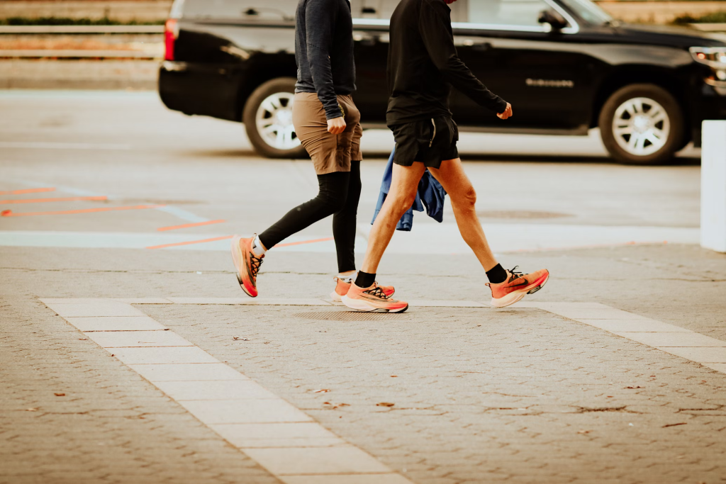 Two people's legs in athletic shoes and leggings walking across concrete pavement near parked cars.