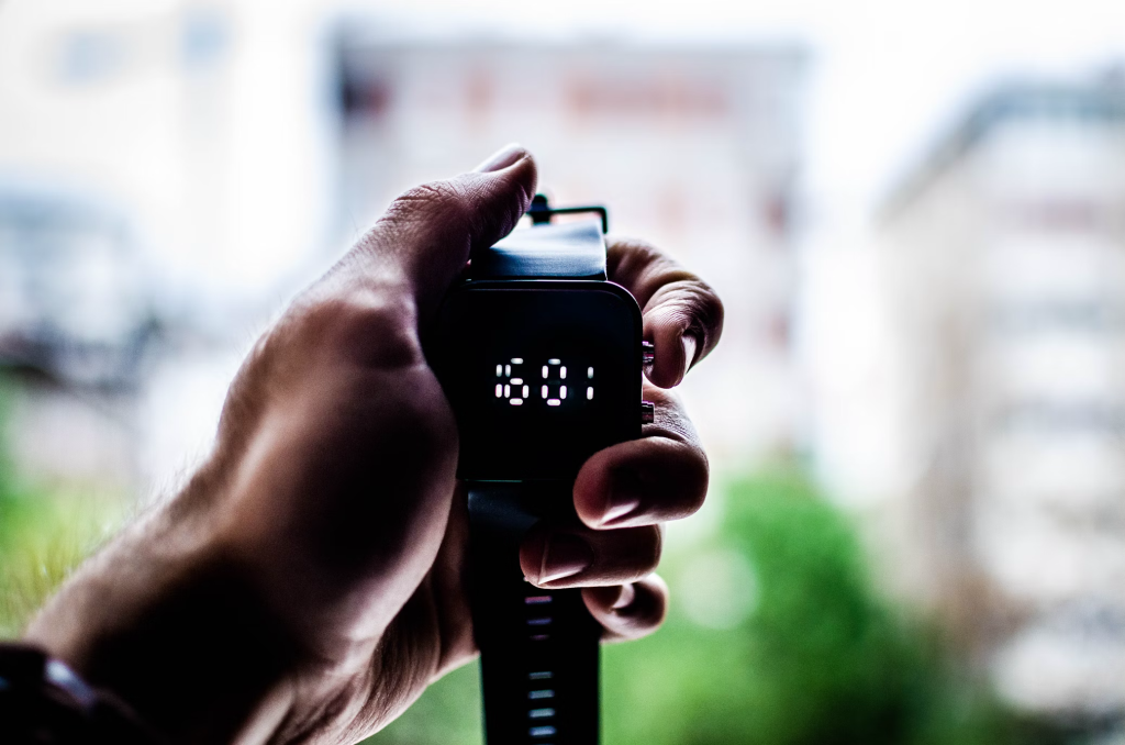 Hand holding a black fitness tracker displaying the time 6:01 on its digital screen.