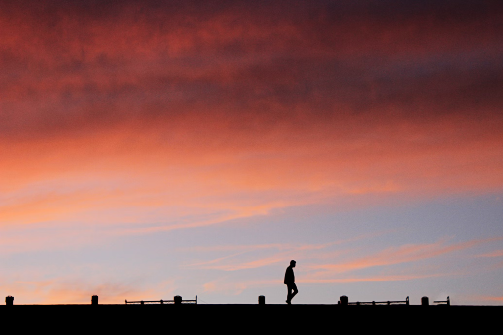 Silhouette of a person walking along a a sidewalk against a dramatic orange and red sunset sky.