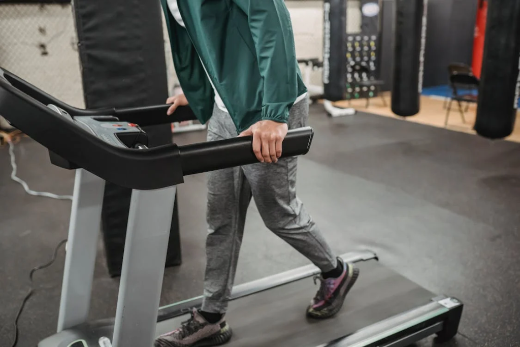 Person in gray athletic wear and green jacket walking on a treadmill in a fitness facility.