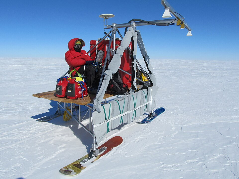 Researchers in red winter gear operating radar equipment on skis across the white Antarctic ice sheet under clear blue skies.