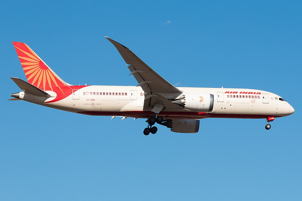 Air India Boeing 787 Dreamliner in flight against blue sky, showing the distinctive red sunburst logo on the tail.