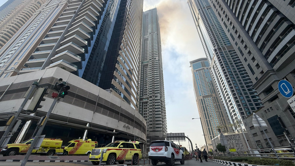 Dubai Civil Defence fire trucks stationed at the base of Marina Pinnacle Tower during emergency response operations, with the towering residential buildings of Dubai Marina visible above.