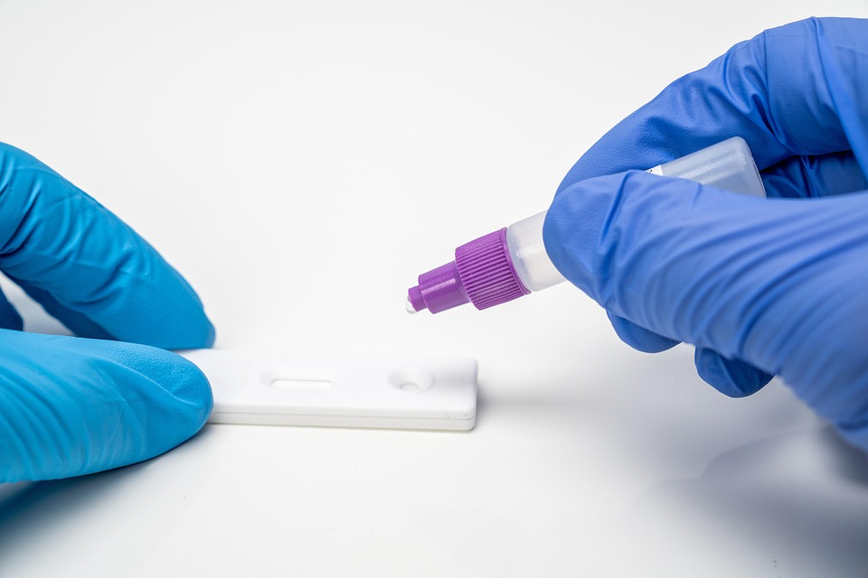 Gloved hands applying a sample to a white COVID-19 rapid antigen test device, showing the type of testing that can quickly confirm infection with the new variant causing razor blade throat symptoms.