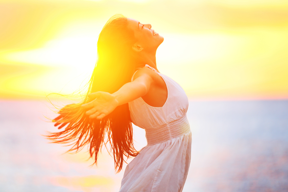 Enjoyment - free happy woman enjoying sunset. Beautiful woman in white dress embracing the golden sunshine glow of sunset with arms outspread and face raised in sky enjoying peace, serenity in nature