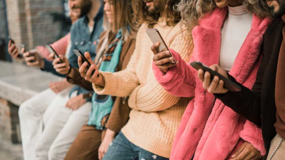 Group of young multi-ethnic people using smartphone sitting in a bench. Multiracial people holding smart mobile phones and watching their screen outdoors.