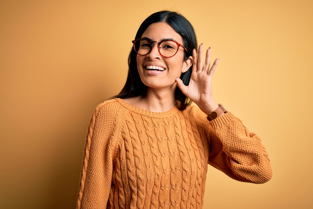 Young beautiful hispanic woman wearing glasses over yellow isolated background smiling with hand over ear listening an hearing to rumor or gossip. Deafness concept.