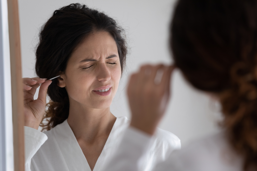 Unhappy young woman do daily facial routine procedures in bathroom at home clean ears with cotton bud or stick. Millennial female look in mirror in shower feel pain take care of body hygiene in bath.