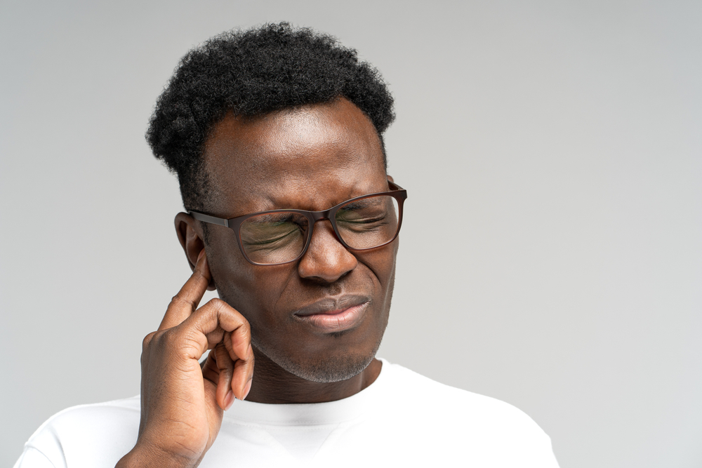 Sick African American millennial man suffering from tinnitus, throbbing earache, tired of noise. Irritated black frowning male in glasses touching painful ear, isolated on grey studio background.