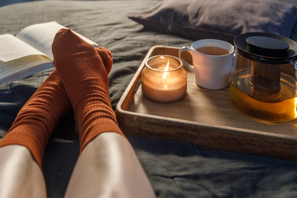 Soft photo of a woman`s legs in orange woolen socks on the bed with a book and cup of tea and a candle on the tray. Interior and home coziness concept. Top view
