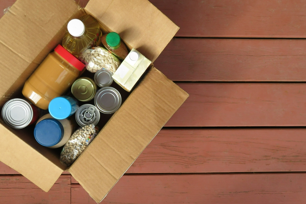 A box of donated canned goods and non-perishable foods for a food pantry for the poor sitting on red, wood panel background. Copy space.