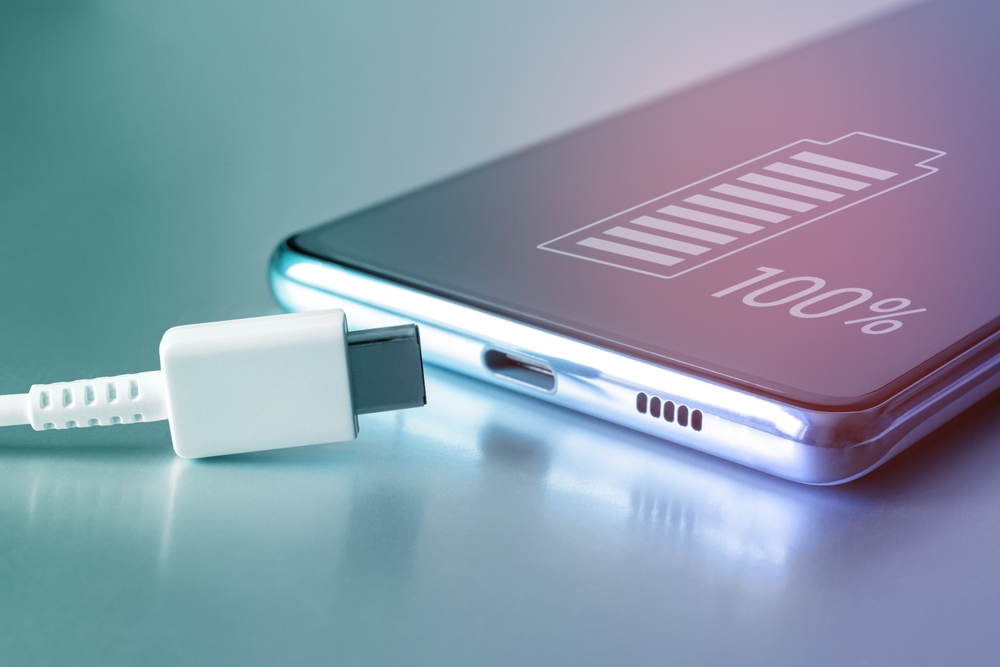 Fully charged battery icon on the touch screen of the cell phone and a USB Type-C cable. Fully charged smartphone battery. Keeping the battery intact. Battery safety. Copy space
