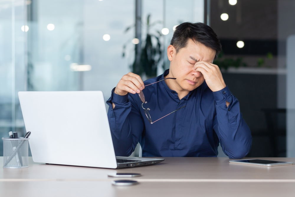 Asian worker tired at work has pain in eyes, businessman in glasses working late, using laptop at work inside office, serious and concentrated pensive.
