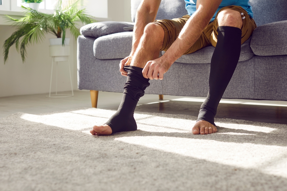 Man wears compression stockings due to varicose or recovery after thrombosis. Unrecognizable man sitting on sofa and covering tired legs with elastic medical anti varicose anti thrombotic sleeve socks