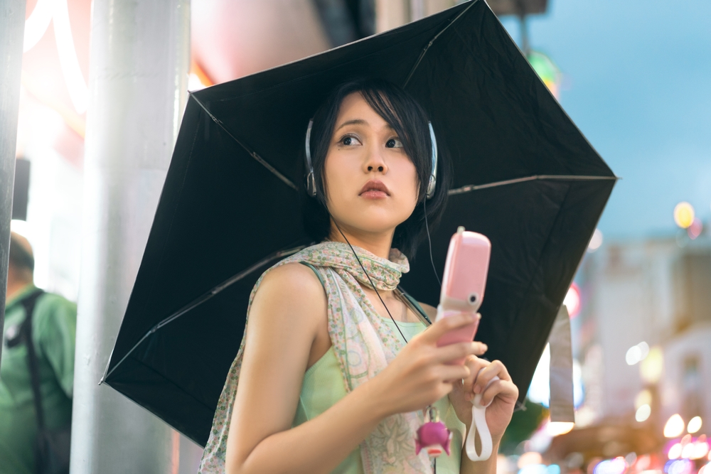 Urban real people lifestyles in downtown concept. Young asian woman wear y2k nostalgia trend outfit stylish using headphone and flip phone. Rainy season with umbrella. Stand in Bangkok city Thailand