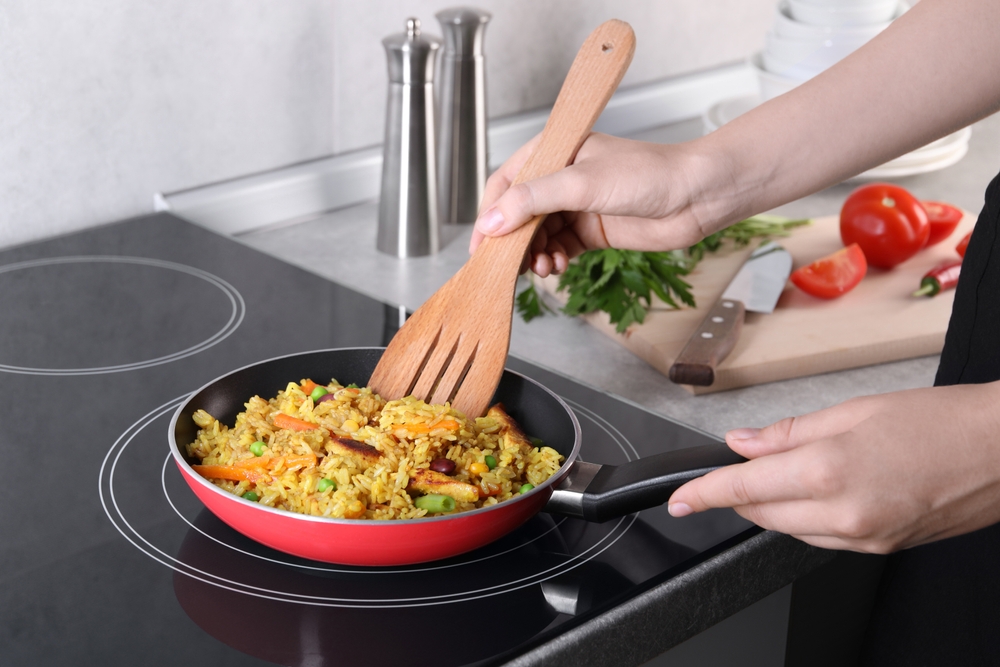 Woman frying rice with meat and vegetables on induction stove in kitchen, closeup