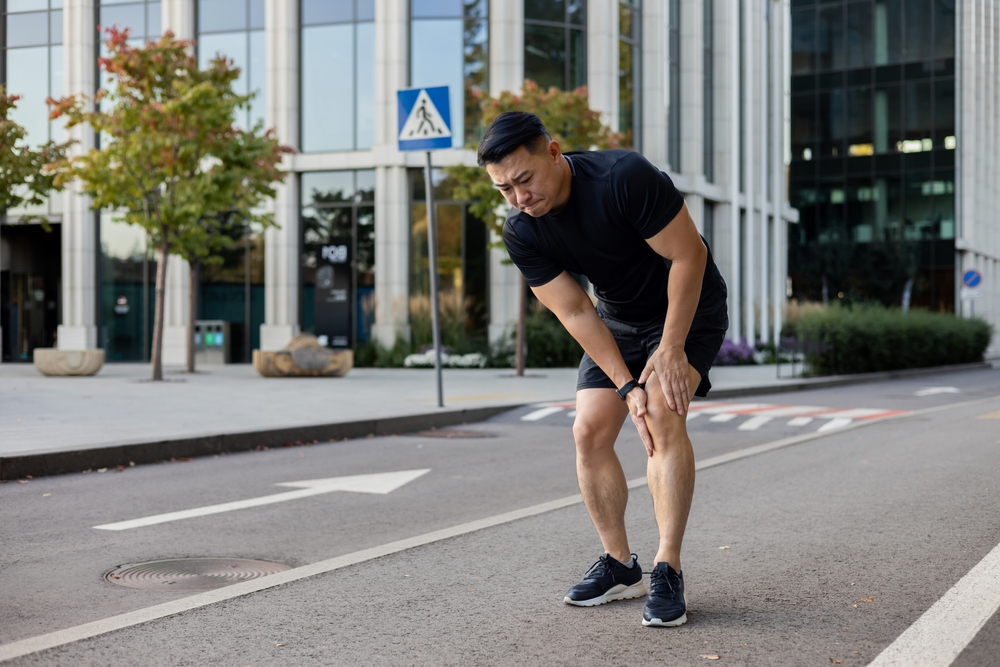 A young Asian man runs in the morning through the streets of the city, stands and holds his leg, feels severe pain and spasm in his knee, has a sports injury.