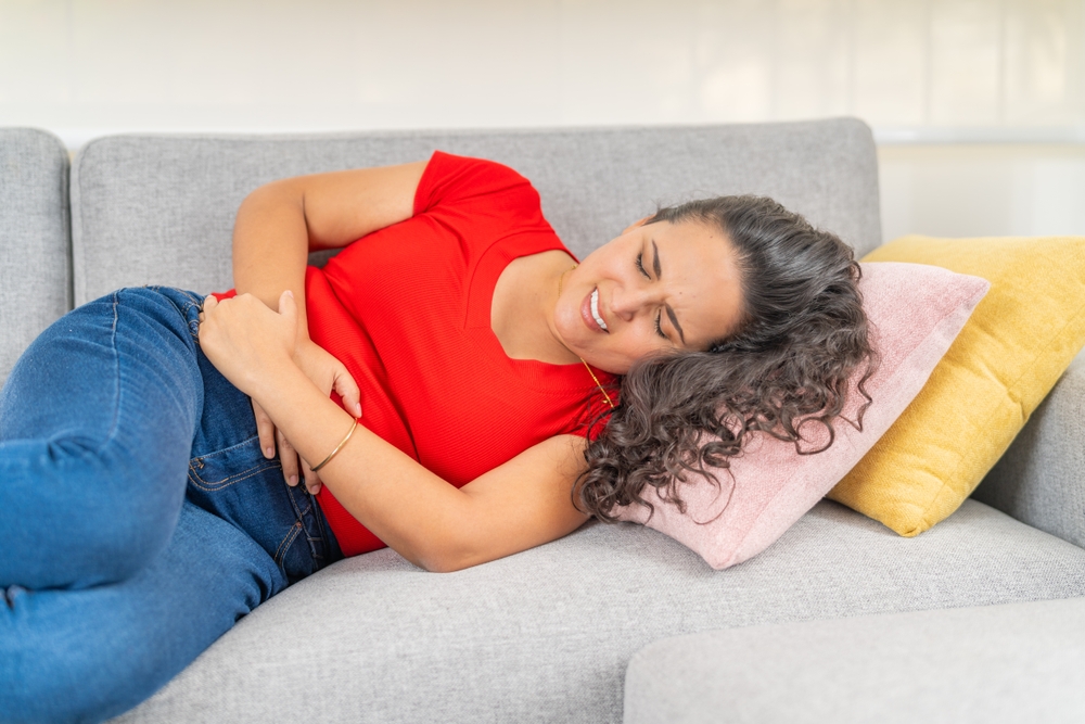Young adult female curled up on a sofa, clutching her stomach in discomfort.
