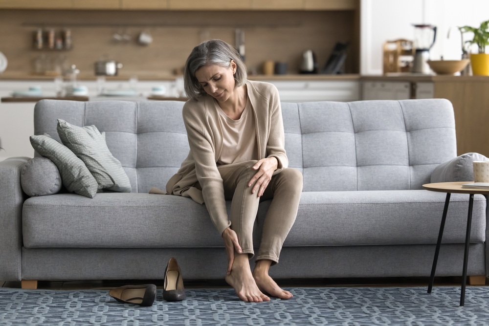 Female came home sit on sofa took off shoes, touch ankle doing self-massage of tired feet, suffers from varicose, painful feelings discomfort after long workday on heels, need rest, orthopedic insoles