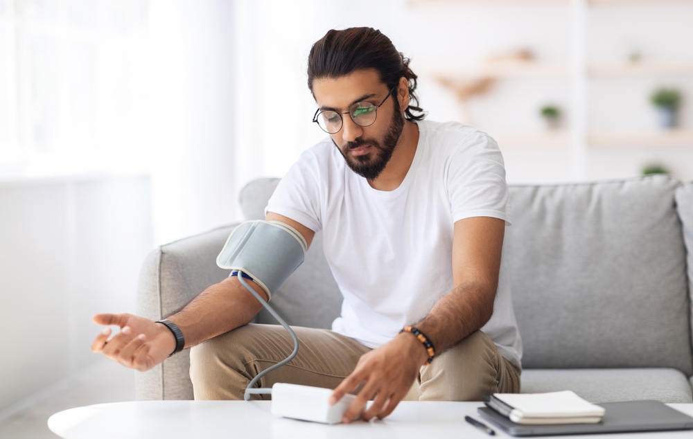 Young arab man sitting on couch, checking blood pressure and taking notes at home. Indian guy in white t-shirt using modern tonometer, experiencing hypertension caused by stress, copy space
