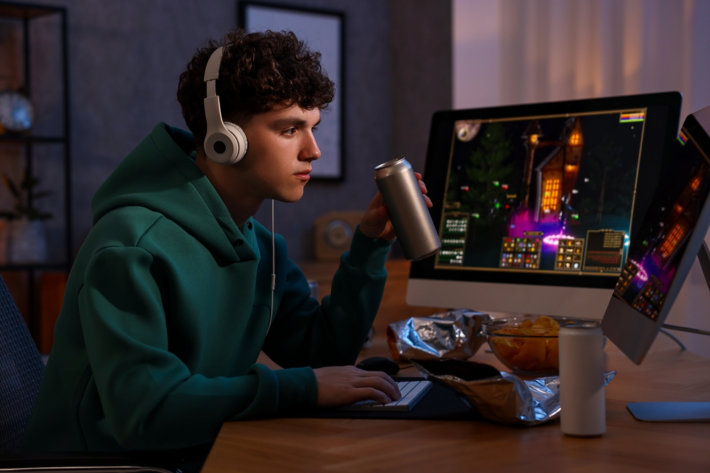 Young man with energy drink and headphones playing video game at wooden desk indoors
