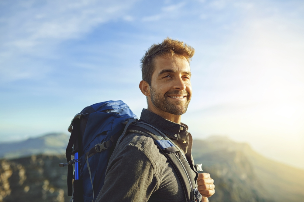 Happy, mountains and hiking with man, backpack and journey with nature, sunshine and getaway trip. Hobby, person and hiker with adventure, travel or environment with wellness, holiday and fitness