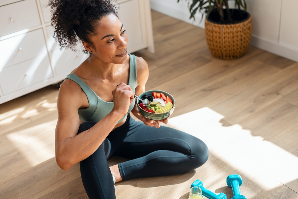 Shot of athletic woman eating a healthy fruit bowl while sitting on floor in the kitchen at home
