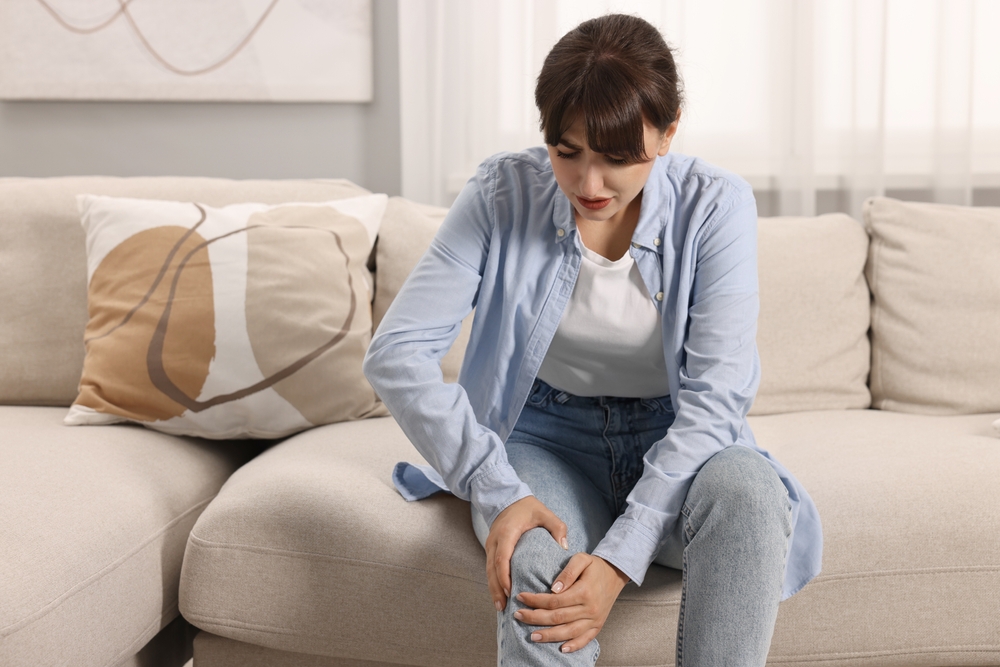 Upset woman suffering from knee pain on sofa at home
