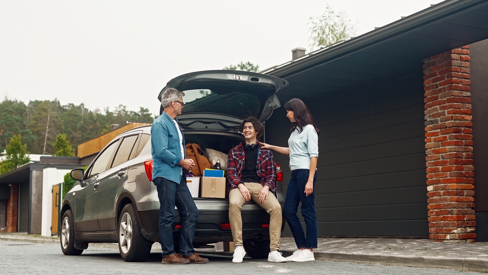 Happy Caucasian parents farewell with their son outside. People standing near grey car full of son's things and communicating with each other. Positively smiling and having good feelings before trip.