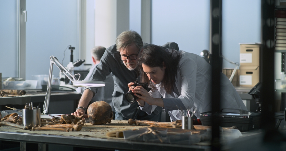 Female paleoanthropologist takes photo of fossil skull remains of extinct human species with colleague archaeologist in archaeological lab using professional camera. Scientist works in the background.
