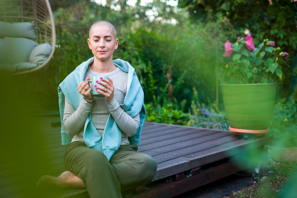 Young female cancer patient sitting on garden patio with coffee, eyes closed. Health insurance, medical expenses, loss of income safety plan. Mental health, wellbeing, meditation during illness.