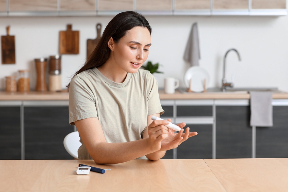 Diabetic young woman using lancet pen at table in kitchen

